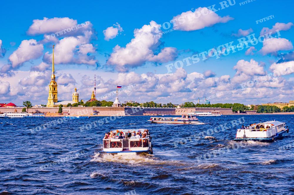 Фотография представляет живописный вид на Петропавловскую крепость в Санкт-Петербурге с реки Невы. На переднем плане видны туристические катера, полные пассажиров, наслаждающихся речной прогулкой по волнам под парусом белоснежных облаков в голубом небе. Это изображение подойдёт для статей о Санкт-Петербурге, туристических проспектах, блогах о путешествиях и как реклама речных круизов по Неве.